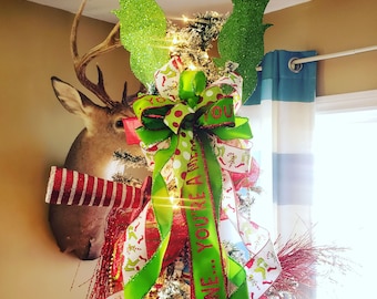 May include: A Christmas tree decorated with Grinch-themed ornaments. The tree features green, glittery Grinch arms and red Santa hats attached to the ceiling. A large green and red bow with the text "You're a mean one" adorns the top.