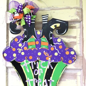 May include: A Halloween-themed wooden door hanger shaped like a cupcake, featuring witch legs and the words "Trick or Treat." The cupcake has a purple top with yellow polka dots and a green base. A decorative bow is attached at the top.