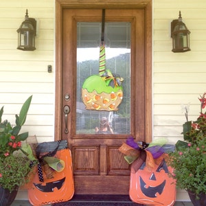 May include: A wooden front door decorated for Halloween with a large green apple-shaped decoration and two orange jack-o'-lantern cutouts. The apple has candy corn accents. The door is flanked by potted plants and lanterns.