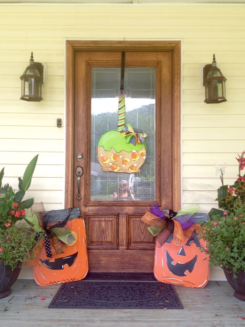 May include: A wooden front door decorated for Halloween with a large green apple-shaped decoration and two orange jack-o'-lantern cutouts. The apple has candy corn accents. The door is flanked by potted plants and lanterns.