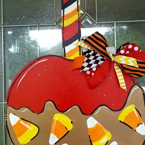 May include: A wooden, apple-shaped door hanger with a red candy apple design. The apple is decorated with brown "caramel" and candy corn. A decorative bow with various patterns and colors is attached to the top. The hanger is painted with stripes.