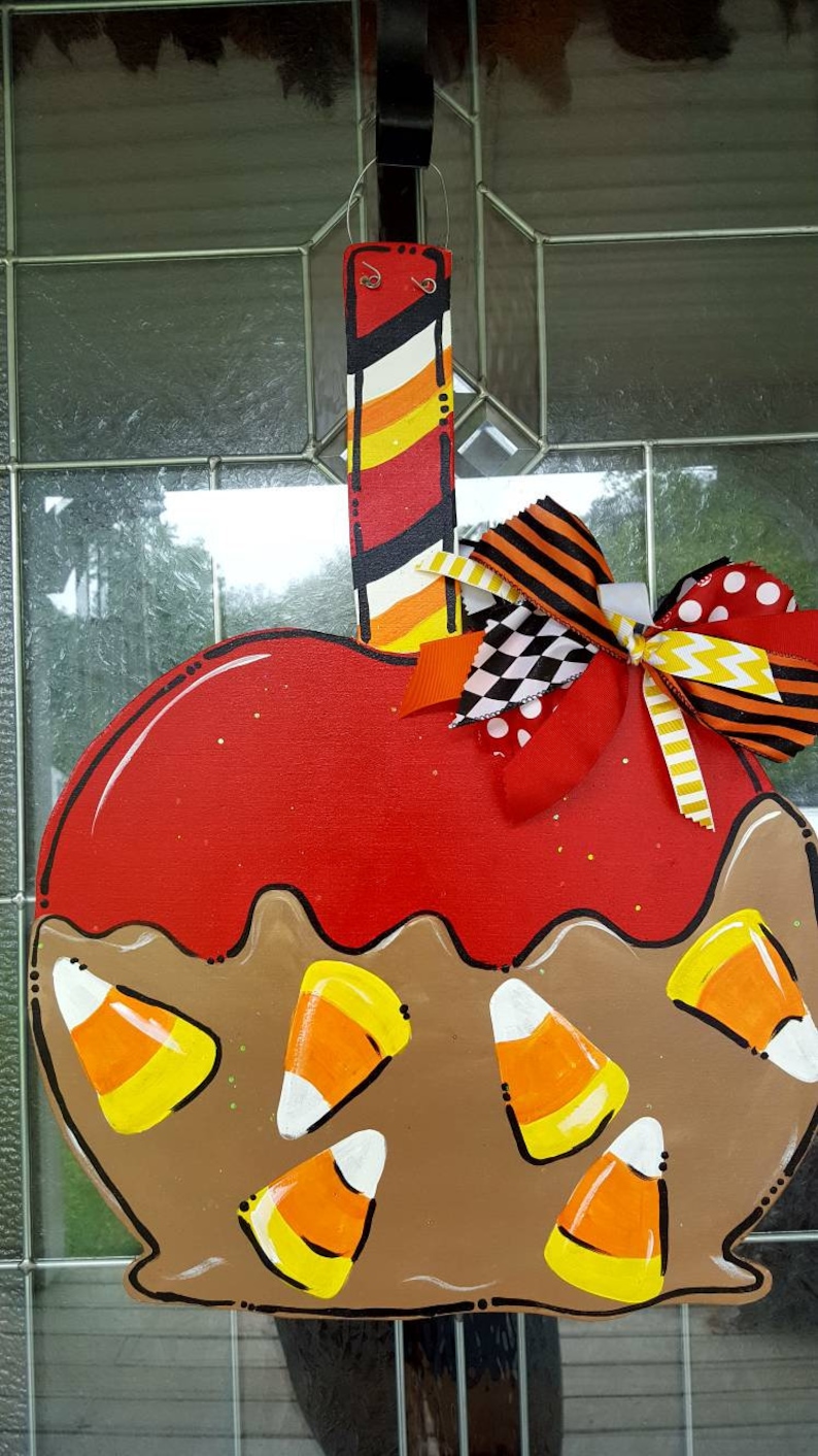 May include: A wooden, apple-shaped door hanger with a red candy apple design. The apple is decorated with brown "caramel" and candy corn. A decorative bow with various patterns and colors is attached to the top. The hanger is painted with stripes.