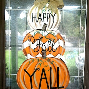 May include: A three-tiered pumpkin-shaped wooden sign with the words "HAPPY", "Fall", and "Y'ALL" in black lettering. The top pumpkin is white, the middle is orange and white, and the bottom is orange. A burlap bow is at the top.