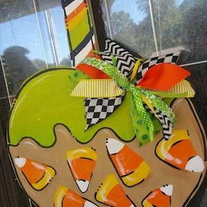 May include: A wooden Halloween-themed apple-shaped decoration. The apple is painted green with a brown base and candy corn accents. A colorful striped stick and a decorative bow with orange, green, and black ribbons complete the design.
