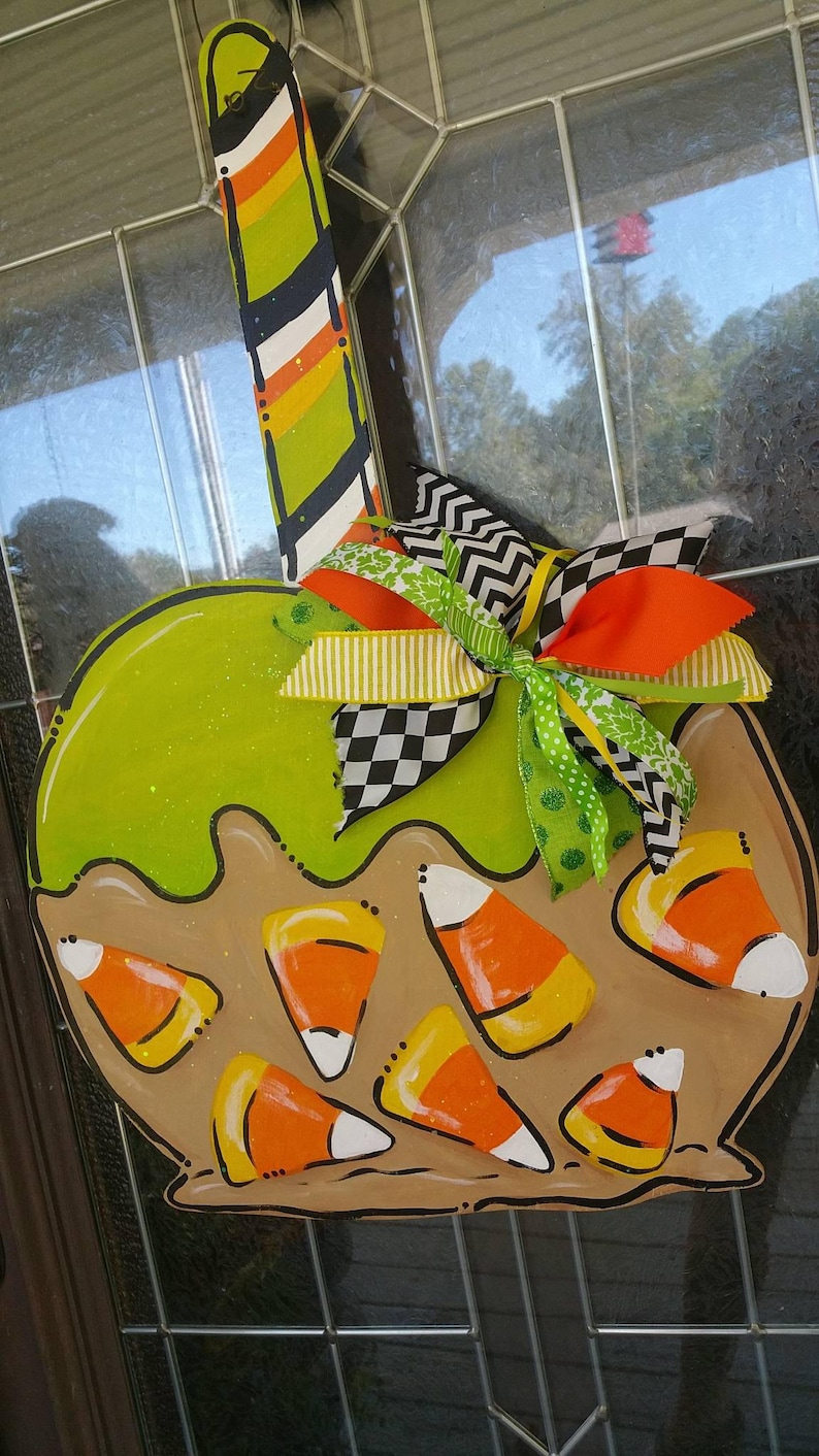 May include: A wooden Halloween-themed apple-shaped decoration. The apple is painted green with a brown base and candy corn accents. A colorful striped stick and a decorative bow with orange, green, and black ribbons complete the design.
