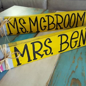 May include: Two wooden pencil-shaped signs with pink erasers and black lettering. The signs read "Ms. McBroom" and "Mrs. Benson".