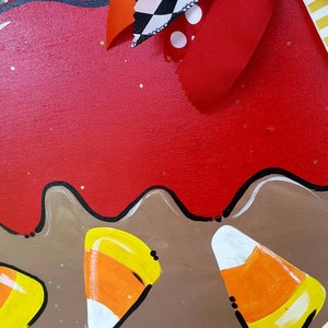 May include: A wooden decoration with a red top and brown base, adorned with painted candy corn. The decoration features a wavy edge and is embellished with colorful ribbons in orange, black, and white patterns, perfect for Halloween.