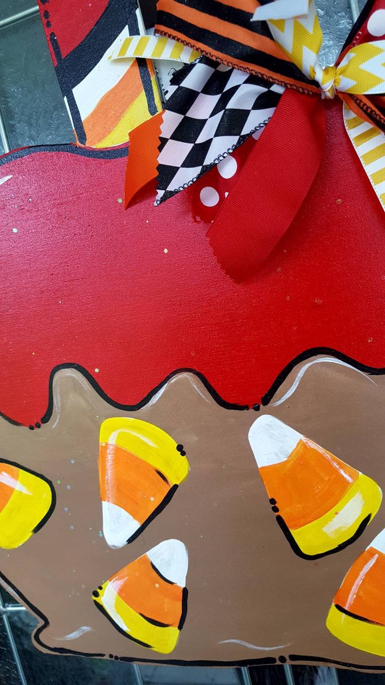 May include: A wooden decoration with a red top and brown base, adorned with painted candy corn. The decoration features a wavy edge and is embellished with colorful ribbons in orange, black, and white patterns, perfect for Halloween.