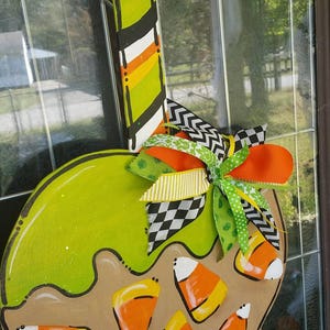 May include: A wooden Halloween-themed door hanger shaped like a candy corn cauldron. The cauldron is green with candy corn accents and a decorative bow. The handle is striped with black, white, orange, and green.