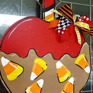 May include: A wooden, apple-shaped Halloween decoration. The apple is painted red with a brown base and candy corn accents. A colorful bow and a striped stick complete the design. The decoration is designed to hang.