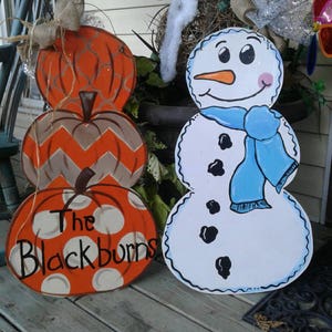 May include: Wooden fall and winter decorations. Three stacked orange pumpkins with various patterns and the text "The Blackburns". A snowman with a blue scarf stands next to the pumpkins. The decorations are on a wooden porch.