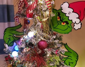 May include: A decorated Christmas tree with white flocking, ornaments, and lights. The tree features Grinch-themed decorations, including cutouts of the Grinch's face and hands. Red and green ribbons and garlands add to the festive holiday decor.