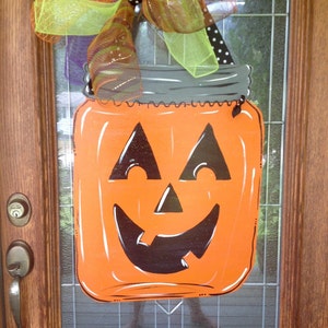 May include: A flat, orange mason jar-shaped wall hanging featuring a black jack-o'-lantern face with triangular eyes and a jagged mouth. The top is decorated with a large, multi-colored mesh bow in lime green, orange, purple, and black polka dots.
