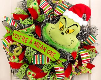 Puede incluir: Una corona navideña con la cara verde del Grinch con un gorro de Papá Noel rojo. La corona está decorada con cintas, lazos a rayas rojas, verdes y blancas, y el texto "YOU'RE A MEAN ONE..."