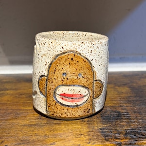 Puede incluir: Una taza cilíndrica de cerámica con un esmalte de color crema moteado. La parte frontal presenta un diseño de cara de mono marrón con dos pequeños ojos oscuros y una boca de color rojo brillante. La taza tiene un borde liso y base plana.