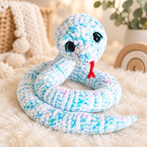 May include: A plush crochet snake toy made of soft white yarn with variegated flecks of light blue, teal, and pink. The snake features large, round, dark green eyes with black pupils and a small, bright red yarn tongue protruding from its mouth.