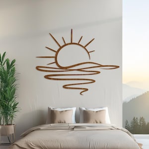 Op de afbeelding: Een bruin, plat, lasergesneden metalen wandkunstwerk dat een gestileerde zon voorstelt die opkomt boven golvende waterlijnen. Het ontwerp toont een halfronde zon met stralen boven drie horizontale, vloeiende, golfachtige lijnen.