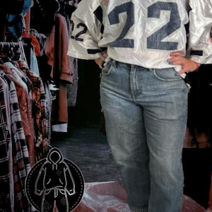 Puede incluir: Una camiseta de fútbol blanca vintage con el número 22 en azul marino. Se combina con vaqueros de mezclilla de lavado claro y zapatillas blancas. El fondo muestra un perchero con ropa.