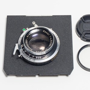 May include: A black Schneider-Kreuznach Xenar 1:4.7/135 lens mounted on a square black lens board with a Compur-P shutter mechanism. The set includes a black circular lens cap and a thin black metal filter ring, all featuring silver text.