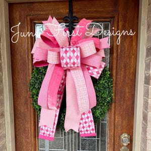 May include: A decorative wreath with a large pink bow, featuring various shades and patterns of pink ribbon. The wreath is hanging on a wooden door with a glass panel. The text "June First Designs" is written on the door.