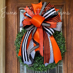 May include: A decorative wreath with a large orange bow, black and white striped and polka dot ribbons. The wreath is made of green foliage and hangs on a wooden door. The text "June First Designs" is visible.