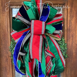May include: A festive wreath with a large bow featuring red, green, and blue ribbons. The bow has a checkered pattern and is accented with gold and plaid ribbons. The wreath is adorned with greenery and is hanging on a wooden door.