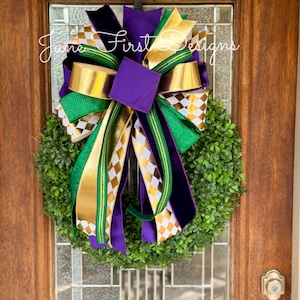 May include: A decorative wreath with a lush green base, adorned with a large bow. The bow features gold, purple, and green ribbons, some with a diamond pattern. The wreath is hanging on a wooden door.
