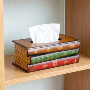 May include: A rectangular tissue box cover designed to resemble a stack of three vintage books. The spines are coloured brown, green, and red, each featuring gold-toned decorative floral and geometric patterns. A white tissue protrudes from the top.