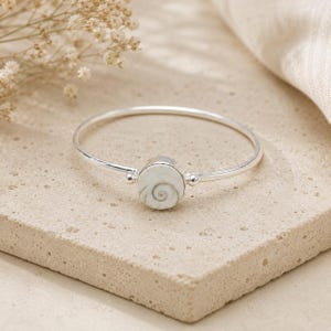 May include: A silver bangle bracelet featuring a circular centre stone with a white and grey spiral pattern. The metal band is thin and polished, connecting to the round setting on both sides with small silver spheres, creating a simple design.