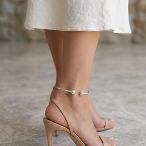 May include: A silver-toned metal anklet featuring a thin, open-ended band design with a polished spherical bead at each end. The anklet is worn around the ankle, positioned above a pair of beige high-heeled sandals with thin straps.