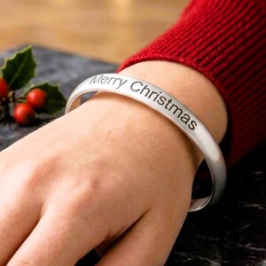 Op de afbeelding: Een zilverkleurige metalen armband met een gladde, matte afwerking. De buitenkant van de armband is gegraveerd met de zwarte tekst "Merry Christmas" in een strak, schreefloos lettertype, die om de pols loopt.