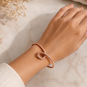 May include: A rose gold-toned metal cuff bracelet featuring a smooth, polished finish and a continuous, looping design that creates a circular knot detail at the center. The bracelet has a rounded profile and an open-ended construction.
