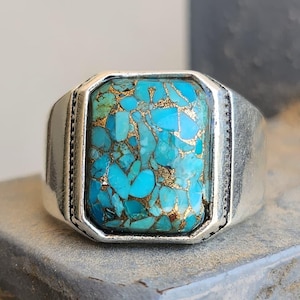 Op de afbeelding: Een zilveren ring met een rechthoekige mozaïek-inleg van helder turquoise steenfragmenten, afgewisseld met metaalachtige goudkleurige aders. De metalen band heeft een gladde, gepolijste afwerking met een subtiele decoratieve rand.