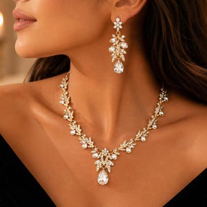 Könnte beinhalten: Ein goldfarbenes Set aus Halskette und Ohrhängern mit klaren, facettierten Kristallen in Blumen- und Blattform. Die Halskette verfügt über einen zentralen tropfenförmigen Kristallanhänger, während die Ohrringe das Design widerspiegeln.