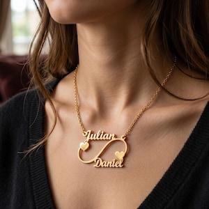 Könnte beinhalten: Eine goldfarbene Halskette mit einem Unendlichkeitszeichen-Anhänger, in den die Namen Julian und Daniel in Schreibschrift eingearbeitet sind. Zwei kleine Herzen sind in das Design der Unendlichkeitsschleife integriert, die an einer feinen Goldkette befestigt ist.