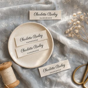 Könnte beinhalten: Vier rechteckige, cremefarbene Stoffetiketten mit dem schwarz aufgedruckten Text Charlotte Bailey und dem Wort HANDMADE in einer kleineren serifenlosen Schrift. Die Etiketten sind auf und um eine kleine, runde, gesprenkelte Schale angeordnet.