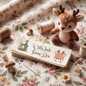 Puede incluir: Una pila de etiquetas de tela rectangulares con el texto With love from Bre, una pequeña ilustración de un hada en una máquina de coser y un ciervo. Las etiquetas están rodeadas de carretes de hilo de madera, una aguja y un peluche.