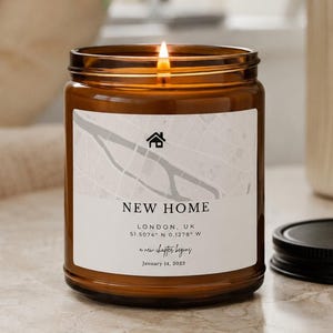 Peut inclure: Une bougie allumée dans un pot en verre ambré avec une étiquette blanche présentant une carte grise, une icône de maison et le texte : NEW HOME, LONDON, UK, 51.5074 N 0.1278 W, a new chapter begins, January 14, 2023. Couvercle noir à côté.