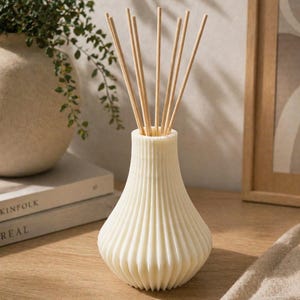 May include: A white reed diffuser with a ribbed, pear-shaped base and a narrow neck. Several light-colored wooden reeds are inserted into the top opening of the vessel, extending upwards in a fan-like arrangement from the center of the container.