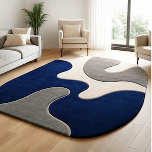 Op de afbeelding: Een vloerkleed met een abstract, organisch patroon met grote, vloeiende vormen in diepblauw, lichtcrème en middengrijs. Het kleed heeft een zachte, pluche textuur en afgeronde randen op de lichtgekleurde houten vloer.