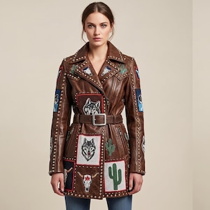 Puede incluir: Una chaqueta de cuero marrón con cuello de solapa, cinturón en la cintura y tachuelas metálicas plateadas a lo largo de las costuras. La chaqueta está decorada con parches bordados de cabezas de lobo, cactus y cráneos de animales con cuernos.