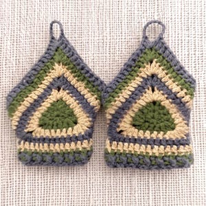 Può includere: Due presine all'uncinetto fatte a mano. Ogni presina presenta un design triangolare con un anello per appenderla. La combinazione di colori include sfumature di verde, beige e grigio, creando un'estetica rustica e accogliente. Per la cucina.