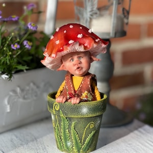 May include: A small figurine of a creature with a large red mushroom cap featuring white spots, wearing a brown vest over a yellow shirt, sitting inside a green textured flower pot decorated with raised green fern leaf patterns.