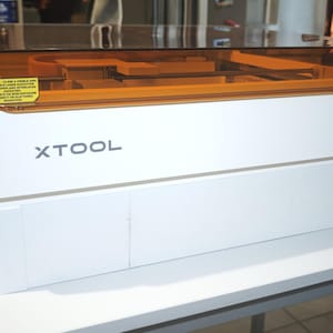 Może przedstawiać: Biała maszyna laserowa xTool z prostokątną, bursztynową, przezroczystą pokrywą. Na przednim panelu znajduje się nazwa marki xTool w kolorze ciemnoszarym oraz żółta etykieta ostrzegawcza dotycząca niewidzialnego promieniowania laserowego.
