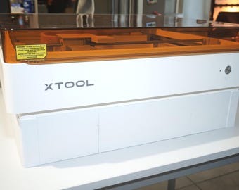 Puede incluir: Una máquina láser xTool blanca con una tapa rectangular transparente de color ámbar. El panel frontal muestra el nombre de la marca xTool en letras gris oscuro e incluye una etiqueta de advertencia amarilla sobre radiación láser.