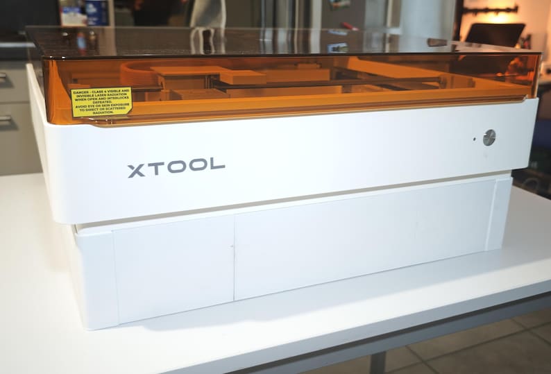 Puede incluir: Una m&aacute;quina l&aacute;ser xTool blanca con una tapa rectangular transparente de color &aacute;mbar. El panel frontal muestra el nombre de la marca xTool en letras gris oscuro e incluye una etiqueta de advertencia amarilla sobre radiaci&oacute;n l&aacute;ser.