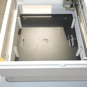 Puede incluir: Una m&aacute;quina de corte l&aacute;ser xTool blanca con una superficie de trabajo interior negra y cuatro pilares de soporte blancos. La placa base negra muestra el texto blanco xTool e incluye una marca de cruz central para el alineamiento.