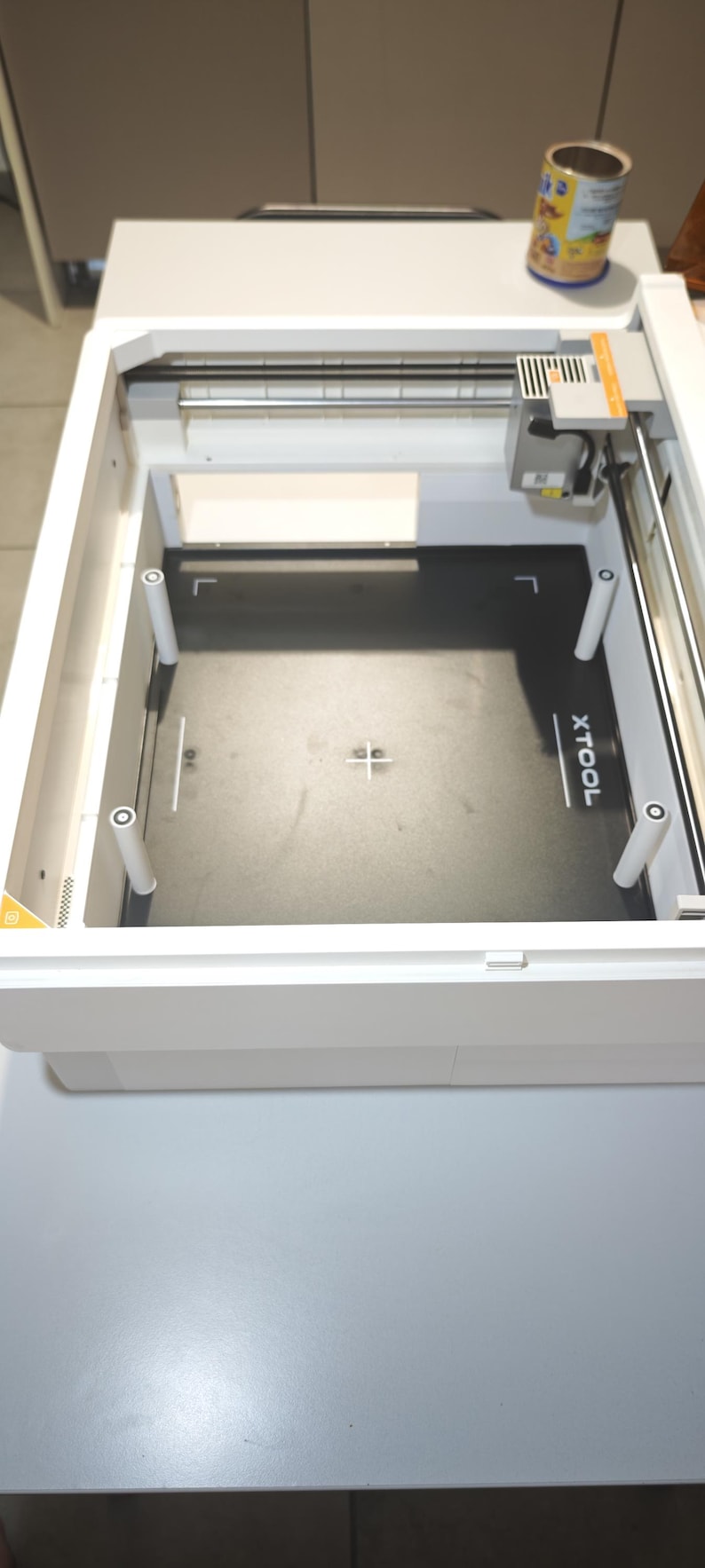 Puede incluir: Una m&aacute;quina de corte l&aacute;ser xTool blanca con una superficie de trabajo interior negra y cuatro pilares de soporte blancos. La placa base negra muestra el texto blanco xTool e incluye una marca de cruz central para el alineamiento.