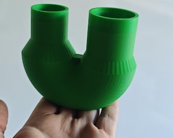 Puede incluir: Un objeto en forma de U de color verde brillante con dos aberturas cilíndricas verticales en la parte superior. La base es redondeada y curva, con un patrón estriado y texturizado en la sección media donde los dos tubos se unen al fondo.