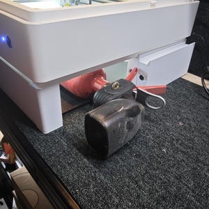 Puede incluir: Una cortadora l&aacute;ser blanca con tapa transparente naranja se sit&uacute;a sobre un extintor rojo y un cencerro negro. La m&aacute;quina cuenta con una base rectangular blanca con una luz indicadora de encendido azul en el panel lateral delantero izquierdo.